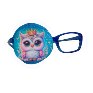 May include: A blue eye patch featuring a colourful cartoon owl wearing a crown and surrounded by flowers. The eye patch is attached to a pair of blue framed glasses.