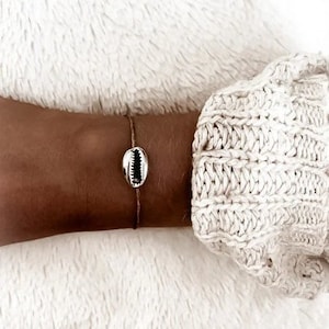 May include: A delicate bracelet with a small, white seashell charm. The bracelet is made of a thin, brown cord and is worn on a person's wrist.