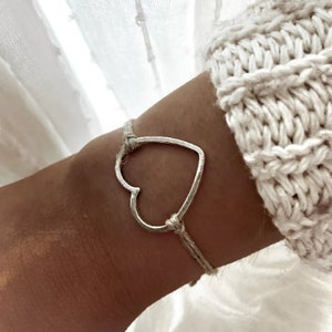 Peut inclure: Un bracelet simple avec un charm en forme de cœur argenté et une ficelle blanche. Le bracelet est porté au poignet.