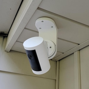 May include: A white security camera mounted on a white bracket, attached to a light-colored exterior wall. The camera has a black front panel and is cylindrical in shape. The bracket is curved and has screw holes.