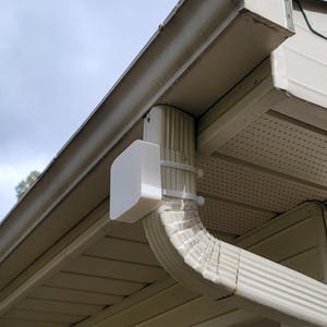 May include: A white, rectangular device is mounted on a white gutter downspout. The device is secured with metal brackets. The downspout is attached to a beige-colored house with a light blue sky in the background. The image is taken from a low angle.