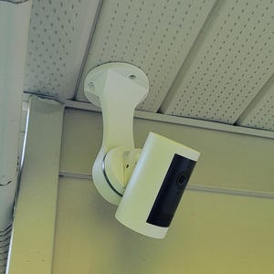 May include: A white security camera mounted on a wall, with a black rectangular section on the front. The camera is attached to a white bracket and is positioned under a white roof overhang.