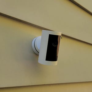 May include: A white security camera with a black front panel is mounted on a beige exterior wall. The camera is cylindrical and attached to a circular base. The wall has horizontal siding and a window is visible in the background.