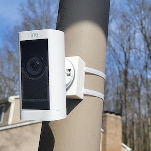 Peut inclure: Une caméra de sécurité Ring blanche montée sur un poteau avec le mot "Ring" imprimé sur la face avant de la caméra.