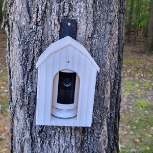 May include: A white, wooden birdhouse-shaped security camera housing mounted on a tree trunk. The camera is black and white, and the housing has a small roof and a rectangular opening. The background shows a forest with green trees and fallen leaves.
