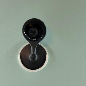 May include: A black Nest security camera mounted on a white circular base against a light green wall.