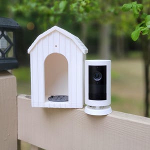 May include: A black and white security camera with a built-in lens sits next to a white dog house-shaped structure. A black lantern sits atop a wooden post. The background is a blurred view of green foliage.