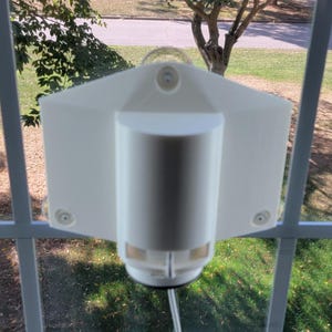 May include: A white, rectangular window cleaning robot attached to a window. The robot has a cylindrical body and two rectangular side panels. The window frame is white, and the background shows a view of a yard and trees.