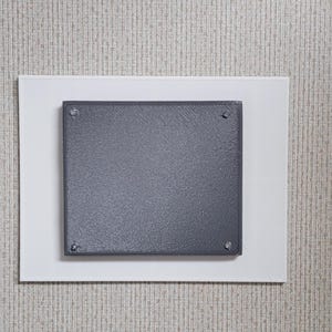 May include: A square, dark gray metal plate is mounted on a white rectangular backing. The plate is secured with four small silver screws. The background is a textured, light beige wall.