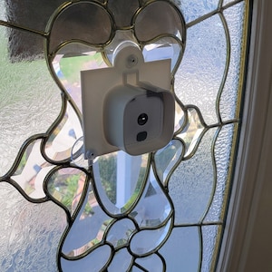 May include: A white security camera mounted on a white plastic bracket, attached to a glass door with a stained glass design. The camera is pointed towards the outside of the door.
