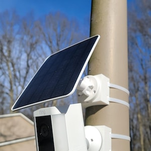 Peut inclure: Une caméra de sécurité blanche avec un panneau solaire noir fixée à un poteau marron. La caméra est fixée avec des sangles blanches et porte la marque "RING". L'arrière-plan présente un ciel bleu et des arbres dénudés.