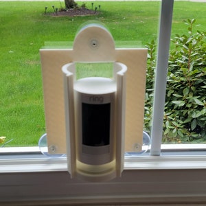 May include: White plastic mount with suction cups attached to a window. A white cylindrical security camera is mounted inside the holder. The camera has a black lens and the text "Ring" is visible on the front.