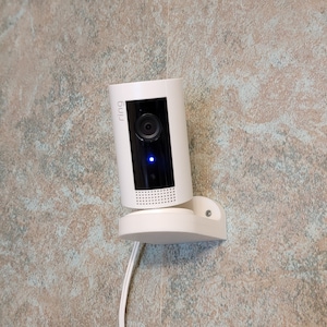 May include: A white security camera mounted on a wall with a white cord.