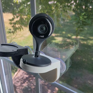 May include: A white security camera with a black lens mounted on a window with a white frame. The camera is pointed towards the outside of the window.
