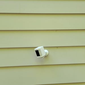 May include: A white security camera mounted on a light yellow siding wall. The camera has a square shape with a black lens and is attached to a round base. A window and door are visible.