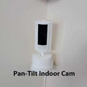May include: A white Pan-Tilt Indoor Cam is mounted in a corner. The camera has a black rectangular lens and is connected to a white base. The text "Pan-Tilt Indoor Cam" is displayed below the camera.