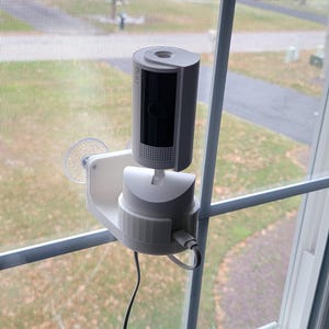 May include: A white security camera mounted on a window. The camera is cylindrical with a black lens and is attached to a white bracket with a suction cup. A black cable extends from the camera.