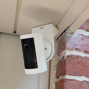 May include: A white security camera with a black front panel is mounted on a white bracket. The camera is attached to a white surface and positioned near a red brick wall. The camera is designed for home security.