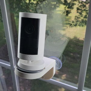 May include: A white Ring security camera mounted on a window. The camera has a black lens and is attached to a white base with a suction cup. The camera is positioned on a window with a view of a green lawn and trees.