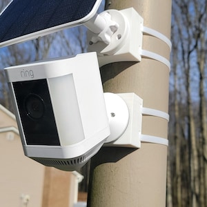 Peut inclure: Une caméra de sécurité Ring blanche avec un panneau solaire fixé à un poteau. La caméra est montée sur un support blanc et possède un objectif noir. La caméra est étiquetée "Ring" sur le devant.