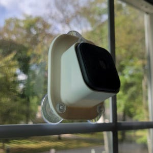 May include: A white and black security camera mounted on a window with a suction cup. The camera is in a white frame and has a black lens. The background shows trees and a blue sky.
