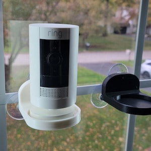 May include: A white Ring security camera with a black front panel is mounted on a window with a suction cup. A black accessory tray is also attached to the window with a suction cup. The camera is cylindrical.