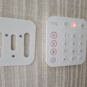 May include: A white Ring Alarm keypad with a red light on, a white mounting plate with three screw holes, and the text "ring" on the keypad.