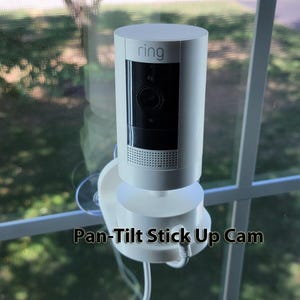 May include: A white Ring Pan-Tilt Stick Up Cam mounted on a window. The camera has a cylindrical design with a black lens and the Ring logo. The text "Pan-Tilt Stick Up Cam" is printed below the camera.