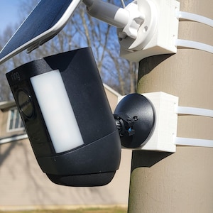 Peut inclure: Une caméra de sécurité noire avec un panneau lumineux blanc est montée sur un poteau avec un panneau solaire fixé sur le côté. La caméra est montée sur un support blanc avec une base noire.