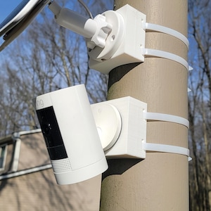 Peut inclure: Une caméra de sécurité blanche montée sur un poteau avec un panneau solaire fixé au sommet. La caméra est montée avec des supports et des sangles en plastique blanc.