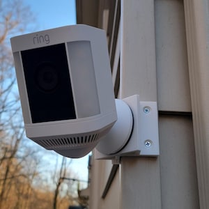 May include: A white Ring security camera mounted on a white siding with a gray background. The camera has a black lens and the word "Ring" is printed on the front.