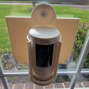 May include: A white Ring security camera mounted on a window with a white plastic cover. The camera is facing forward and the word "ring" is visible on the front of the camera.