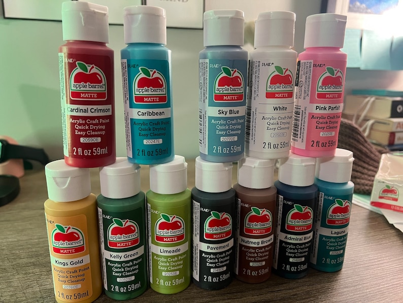 May include: A collection of 10 acrylic craft paint bottles in various colors, including cardinal crimson, caribbean, sky blue, white, pink parfait, kings gold, kelly green, limeade, pavement, nutmeg brown, admiral blue, and laguna. Each bottle is 2 fl oz (59 ml) and features the Apple Barrel brand logo.