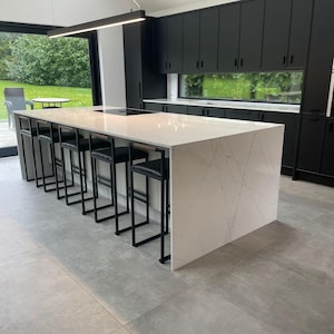 May include: Modern kitchen with a large white countertop island featuring a marble-like pattern. Black metal bar stools with black leather seats are lined up along the island. Black cabinets and a large window are in the background.