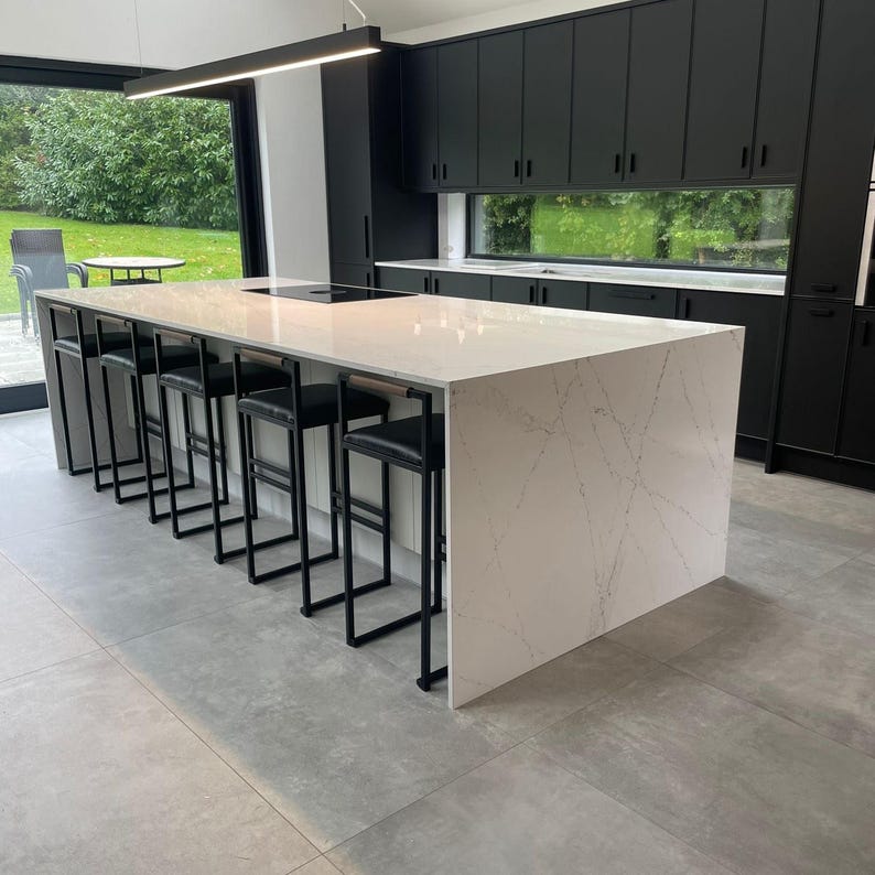 May include: Modern kitchen with a large white countertop island featuring a marble-like pattern. Black metal bar stools with black leather seats are lined up along the island. Black cabinets and a large window are in the background.