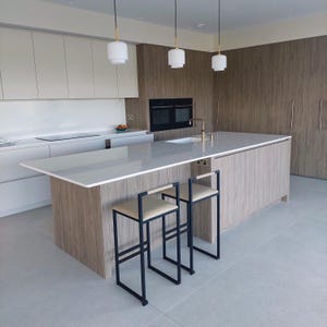 May include: Modern kitchen with light wood cabinetry, a large island with a white countertop, and two black metal bar stools with cream-colored seats. Three pendant lights hang above the island. A built-in oven and sink are also visible.
