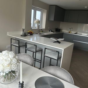 May include: Modern kitchen with a white countertop and gray cabinets. Three gray upholstered bar stools with black metal frames are positioned at the counter. A vase of white roses and a lit candle are on the table in the foreground.
