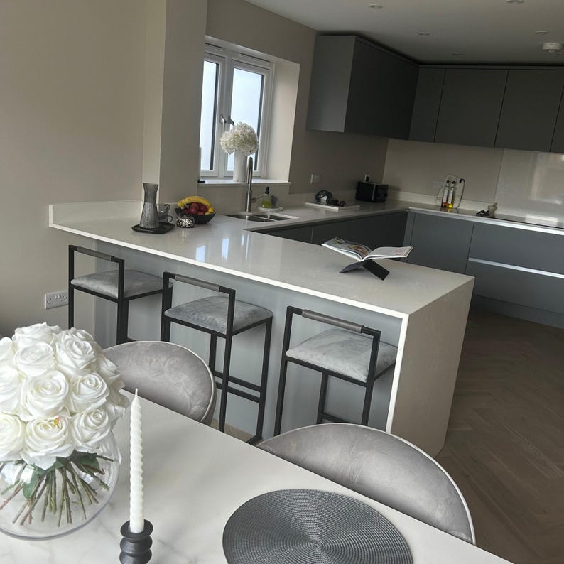 May include: Modern kitchen with a white countertop and gray cabinets. Three gray upholstered bar stools with black metal frames are positioned at the counter. A vase of white roses and a lit candle are on the table in the foreground.