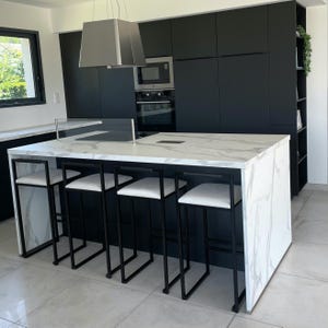 May include: Modern kitchen design featuring a large island with a white marble countertop and black metal bar stools with white seats. The kitchen has black cabinets, a stainless steel range hood, and built-in appliances.
