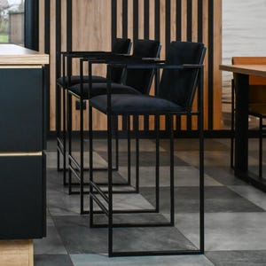 Può includere: Una fila di quattro sgabelli da bar neri con telai metallici quadrati e sedili imbottiti. Gli sgabelli hanno un design minimalista, con poggiapiedi e braccioli. Lo sfondo presenta una parete in legno a strisce verticali e un pavimento a scacchi.