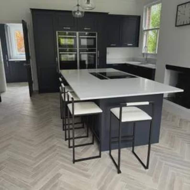 May include: A modern kitchen with dark blue cabinetry and a white countertop island. Four black metal bar stools with white seats are positioned at the island. The floor has a light herringbone pattern.