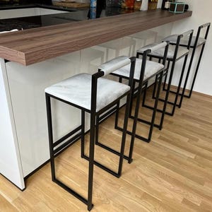 May include: Four modern bar stools with black metal frames and white cushioned seats are lined up against a wooden countertop. The stools have a minimalist design, with a square seat and a backrest. The floor is made of light-colored wood.