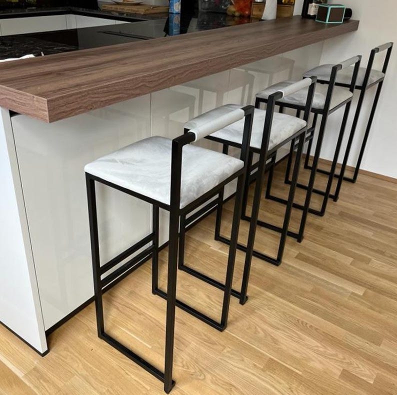May include: Four modern bar stools with black metal frames and white cushioned seats are lined up against a wooden countertop. The stools have a minimalist design, with a square seat and a backrest. The floor is made of light-colored wood.