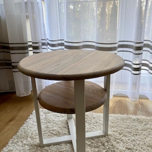 Könnte beinhalten: Ein weißer Holz-Beistelltisch mit runder Tischplatte und einem unteren Regal. Der Tisch hat ein weißes Metallgestell und steht auf einem weißen Shaggy-Teppich.