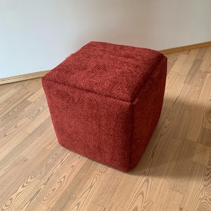 Może przedstawiać: Sześcianowy puf w głębokim czerwonym kolorze. Puf ma teksturowaną, pluszową powierzchnię i stoi na drewnianej podłodze. Design jest prosty i nowoczesny, odpowiedni dla różnych stylów wnętrz.