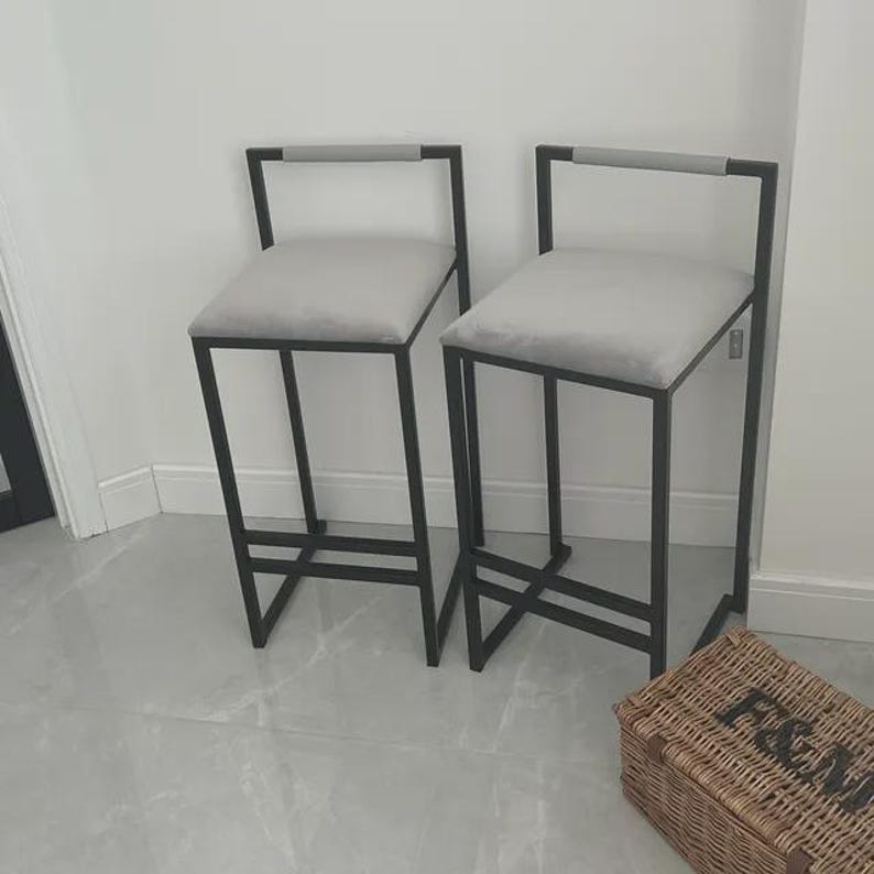 May include: Two modern bar stools with gray upholstered seats and black metal frames. Each stool has a rectangular seat and a backrest with a gray handle. The stools are placed against a white wall.