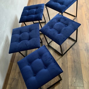 Könnte beinhalten: Vier blaue, quadratische Hocker aus Samt mit schwarzen Metallrahmen. Die Hocker sind versetzt auf einem Holzboden angeordnet.