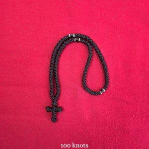 May include: A black knotted cord necklace featuring a cross pendant at the bottom. The necklace is constructed from a continuous cord with a series of intricate knots throughout the length. The text 100 knots is printed in white at the bottom.