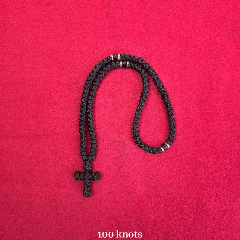 May include: A black knotted cord necklace featuring a cross pendant at the bottom. The necklace is constructed from a continuous cord with a series of intricate knots throughout the length. The text 100 knots is printed in white at the bottom.