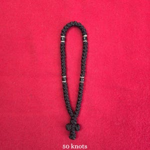 May include: A black knotted cord prayer rope featuring fifty individual knots arranged in a continuous loop. The rope includes small silver-colored spacer beads positioned at intervals and terminates in a knotted cross at the bottom.